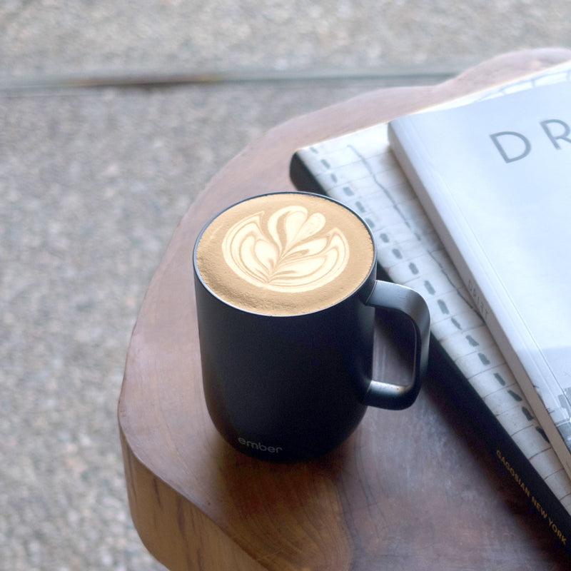 Latte art in a black Ember Mug on a dark wooden table with notebooks..