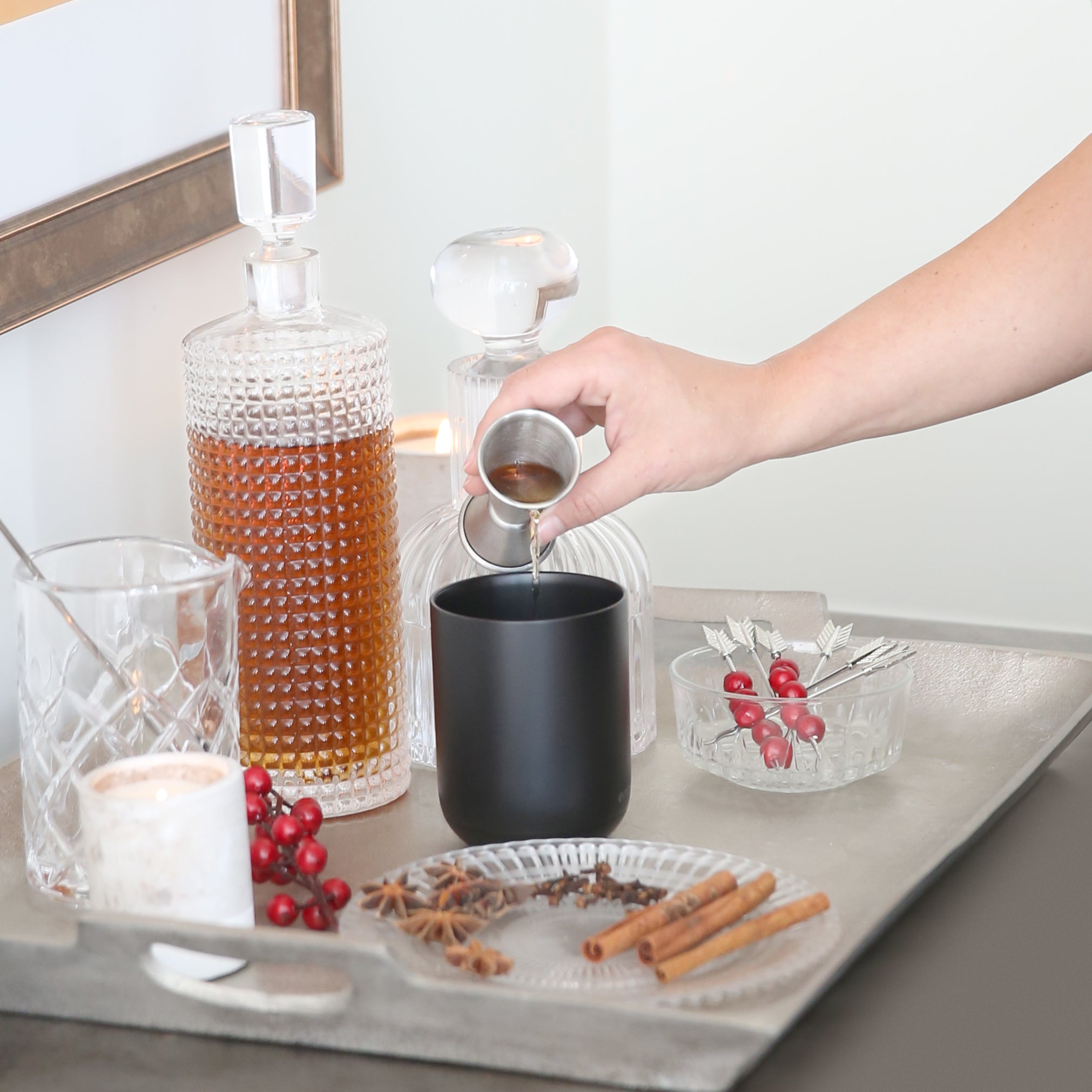 A hand pours liquor into a black Ember Mug² that's surrounded with ornate glassware, cinnamon sticks, cloves, star anise, and cranberries.