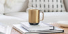 A Gold Ember Mug² sits on top of a white stack of books on a wicker table. There are white cushions and pillows in the background.