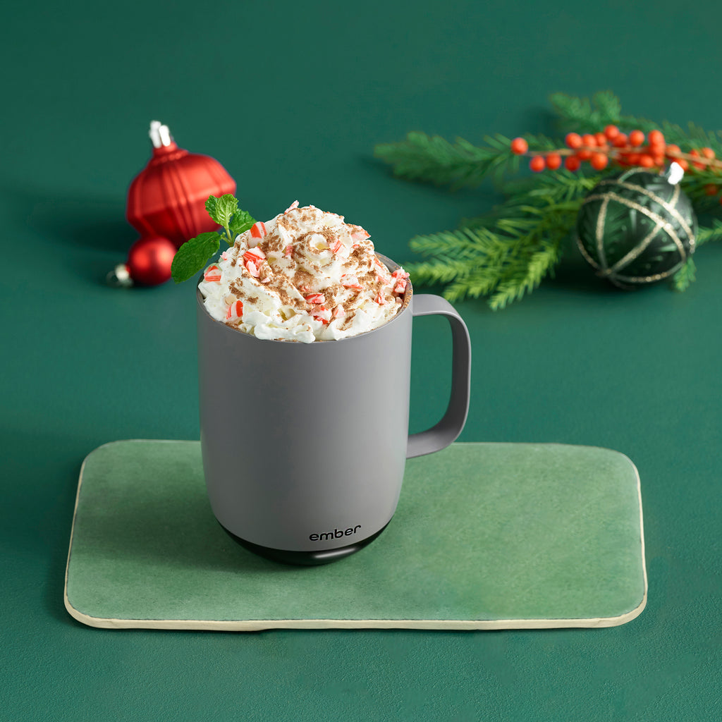 A Gray Ember Mug² is filled with whipped cream and topped with crushed candy cane and cinnamon. The mug sits on a dark green background filled with red and green holiday ornaments alongside a leafy branch with red berries.