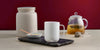 A white Ember Mug² sits on top of a dark marbled cutting board. There is a bowl of sugar to the left and a pot of tea to the right with a tea steeper. The background is dark red.