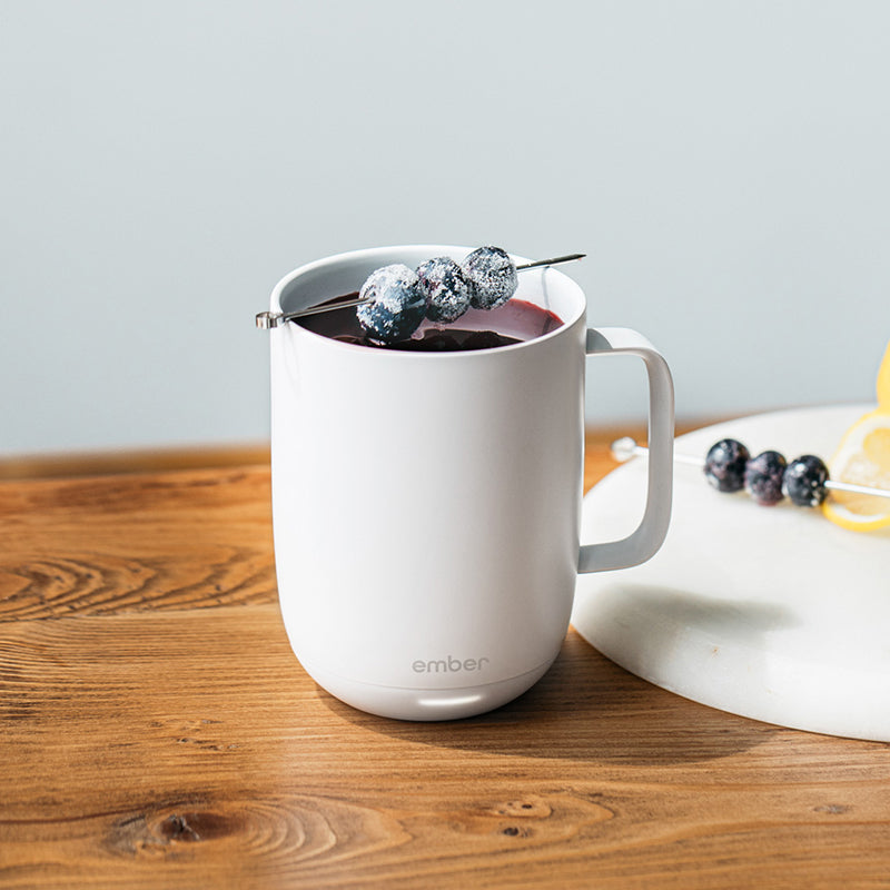 A Gold Ember Mug² filled with black tea sits on a marble and gold table next to a small jar of honey, slices of candied oranges, and a crystal carafe of a brown liquor.